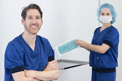 Dr. van Heerbeek en een assistent in blauwe operatiekleding in een klinieksetting. Dr. Lohuis staat met over elkaar geslagen armen en glimlacht, de assistent houdt een gesteriliseerde verpakking met een medisch instrument vast. Ze staan voor een witte muur en een medische tafel.